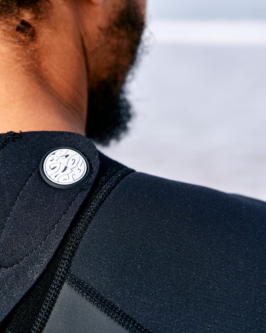Close-up of a person wearing a black neoprene shoulder brace with a white circular logo on the strap.