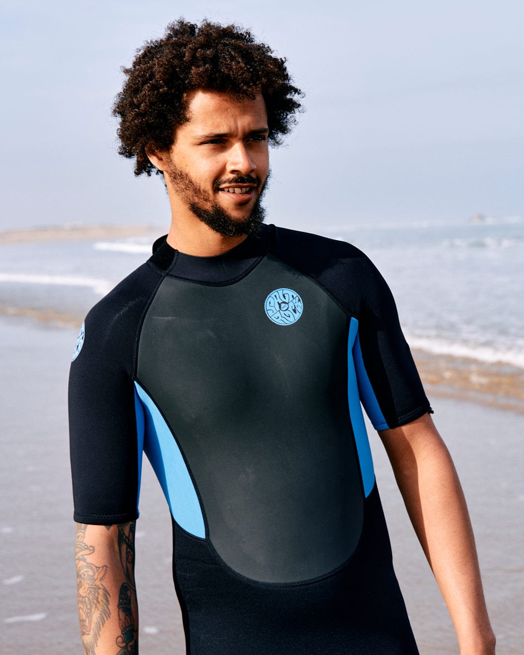 A man with curly hair wearing a Saltrock Core - Men's 3/2 Shortie Wetsuit in Black/blue stands on a beach, looking to the side, with the ocean in the background.