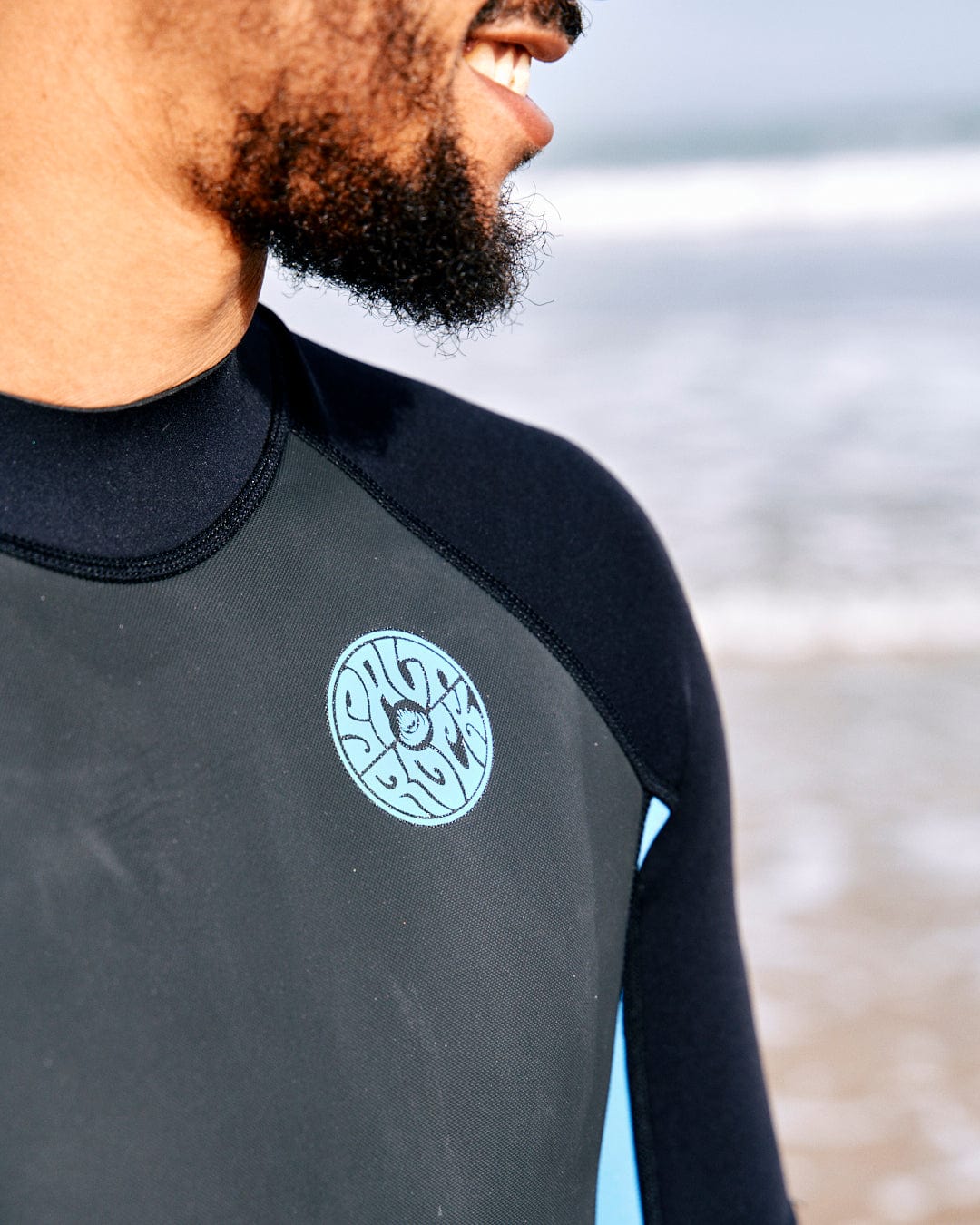 Close-up of a man wearing a Core - Men's 3/2 Shortie Wetsuit in Black/Blue by Saltrock, with a circular logo on the chest, standing by the beach, partially facing the ocean.