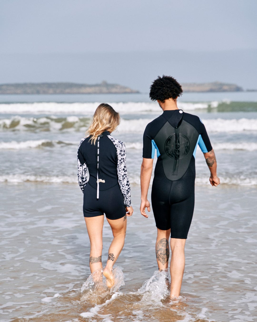Two people in Saltrock neoprene wetsuits walking into the sea, holding hands, with tattoos visible on their arms.