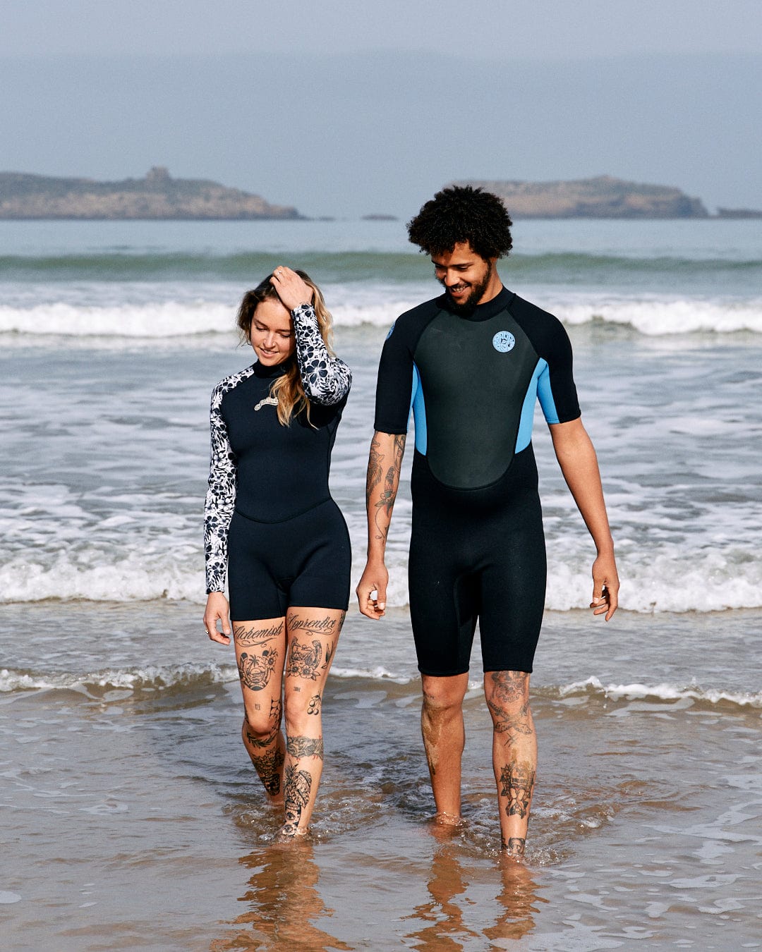 A man and a woman in Saltrock neoprene wetsuits walking hand in hand along a beach, both tattooed, with a clear sky and distant islands in the background.