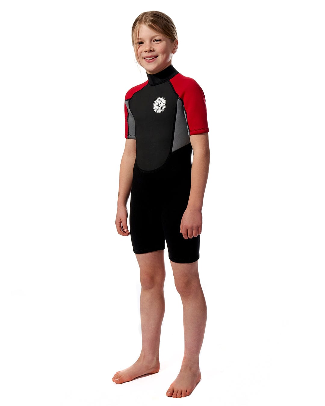 A young girl in a Saltrock Core - Boys 3/2 Shortie Wetsuit in Red stands in front of a white background, ready for beach days.