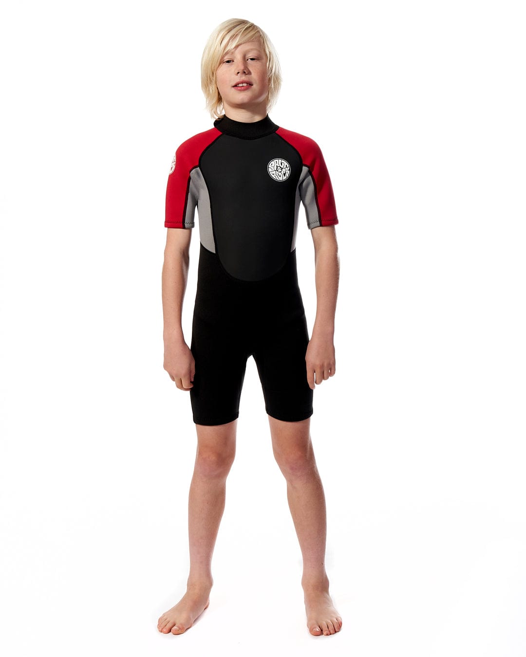 A young boy stands wearing a Saltrock Core - Boys 3/2 Shortie Wetsuit in Black/Red against a white background, looking directly at the camera.