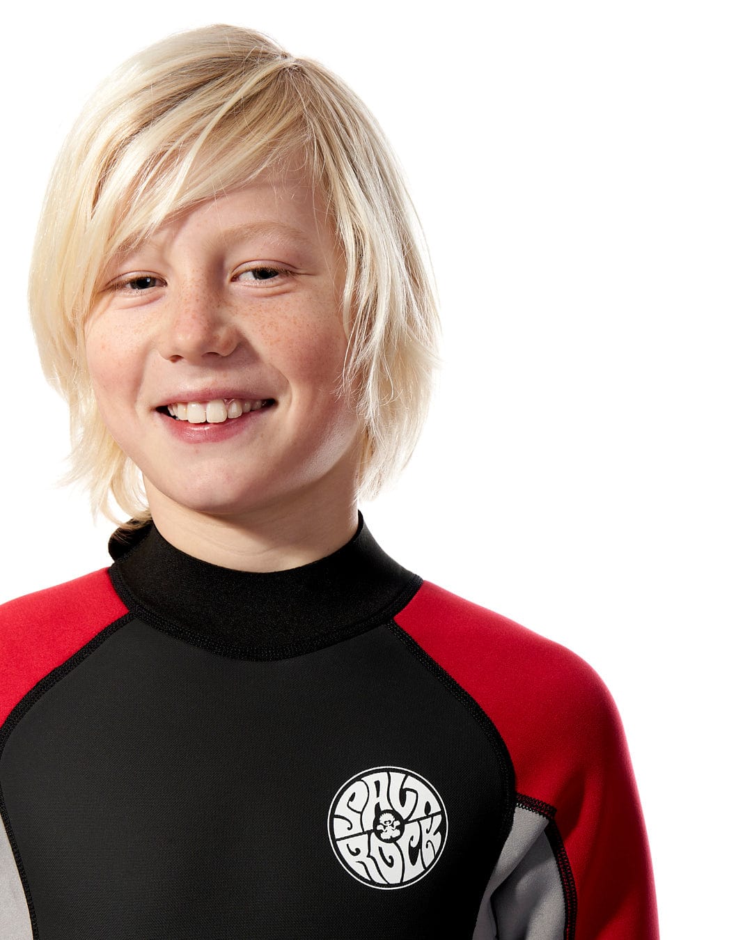 A smiling young boy with blond hair wearing a Saltrock Core - Boys 3/2 Shortie Wetsuit in Black/Red, standing against a white background.