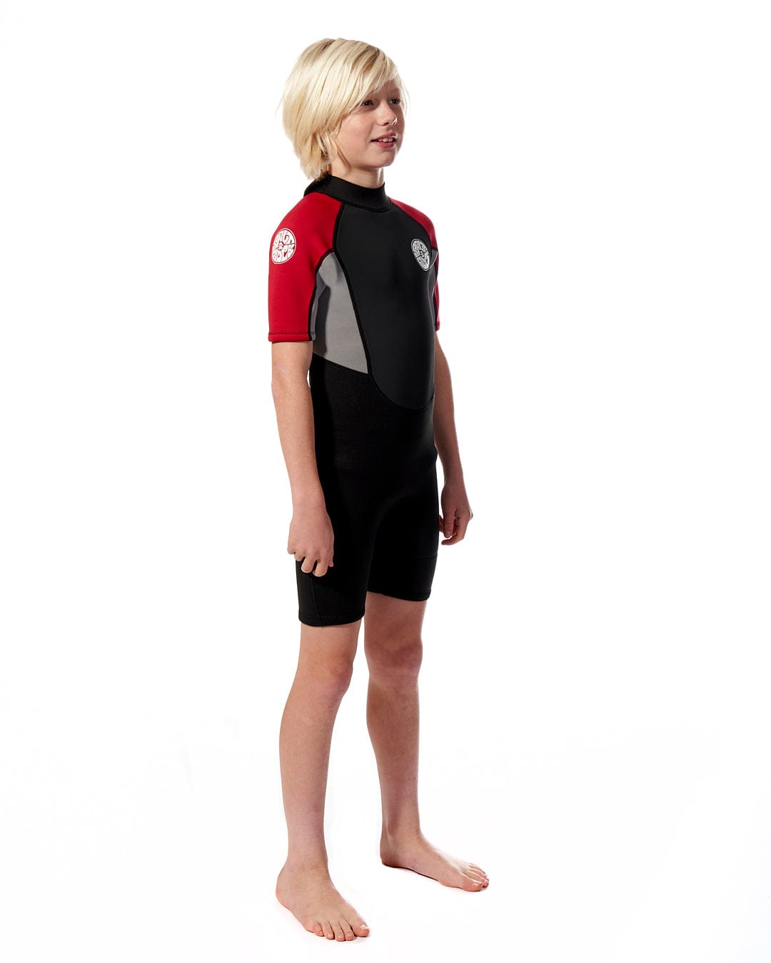 A young boy stands in a Saltrock Core - Boys 3/2 Shortie Wetsuit in black and red against a white background, looking to his left with a slight smile.