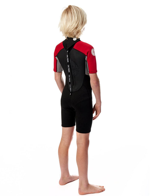 A young boy in a Saltrock Core - Boys 3/2 Shortie Wetsuit - Red at the beach.