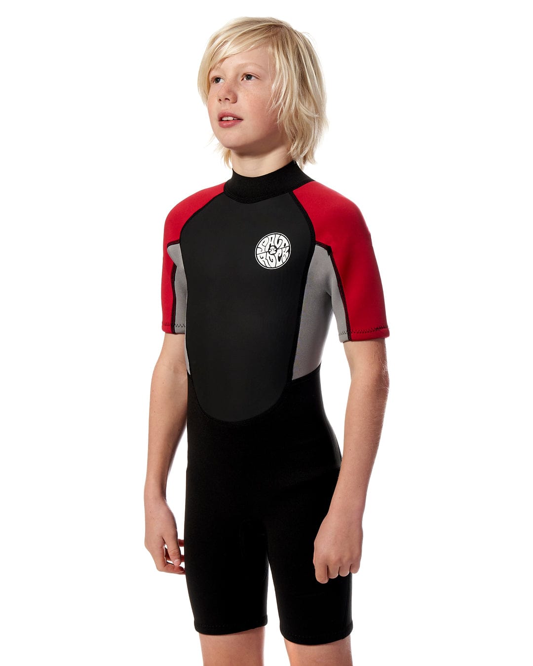 Young boy in a Saltrock Core - Boys 3/2 Shortie Wetsuit - Black/Red standing against a white background. He has tousled blond hair and is looking to the side.
