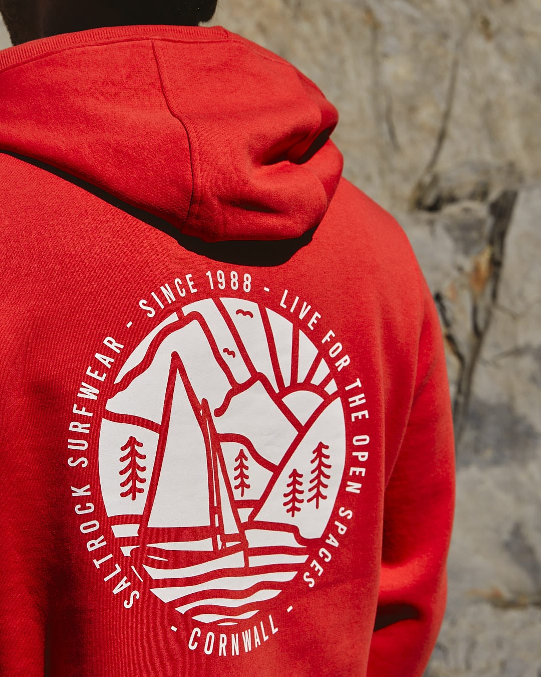 A man wearing a Saltrock Cornwall Sailaway - Mens Pop Hoodie in Red with a sailboat on it.
