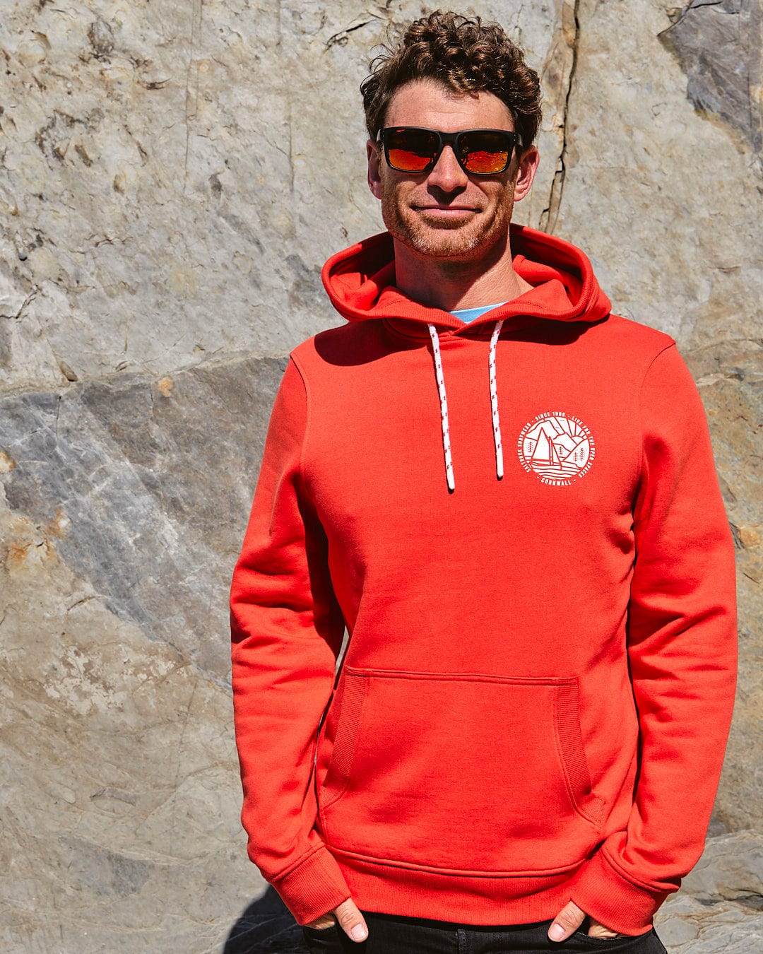 A man in a Saltrock Cornwall Sailaway - Mens Pop Hoodie - Red standing next to a rock.