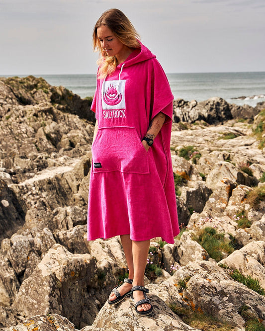 Woman in a pink Saltrock-branded Corp Changing Towel - Bright Pink made of fast-drying cotton towelling material, standing on rocky terrain near the coast.
