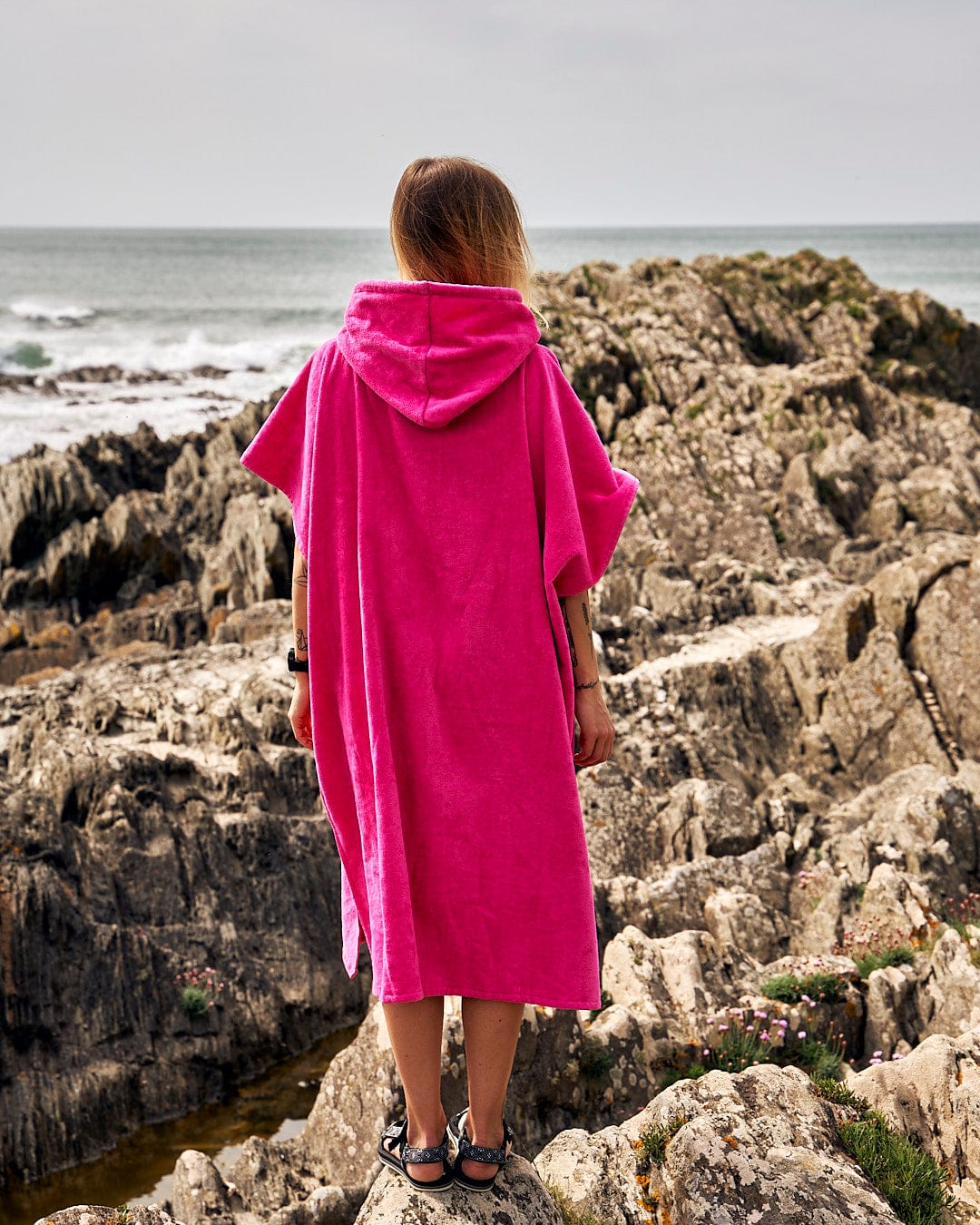 Woman in a Corp Changing Towel - Bright Pink towelling material towel, branded with Saltrock, standing on rocky coast, looking out to sea.