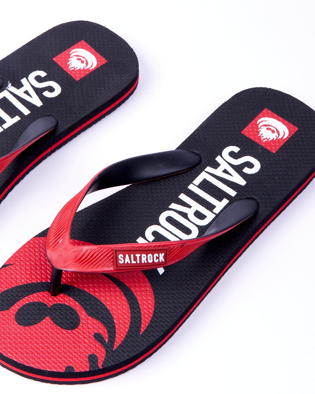 A pair of Saltrock Corp Flame flip-flops with red footbeds featuring a large flamehead logo and black straps, set against a white background.