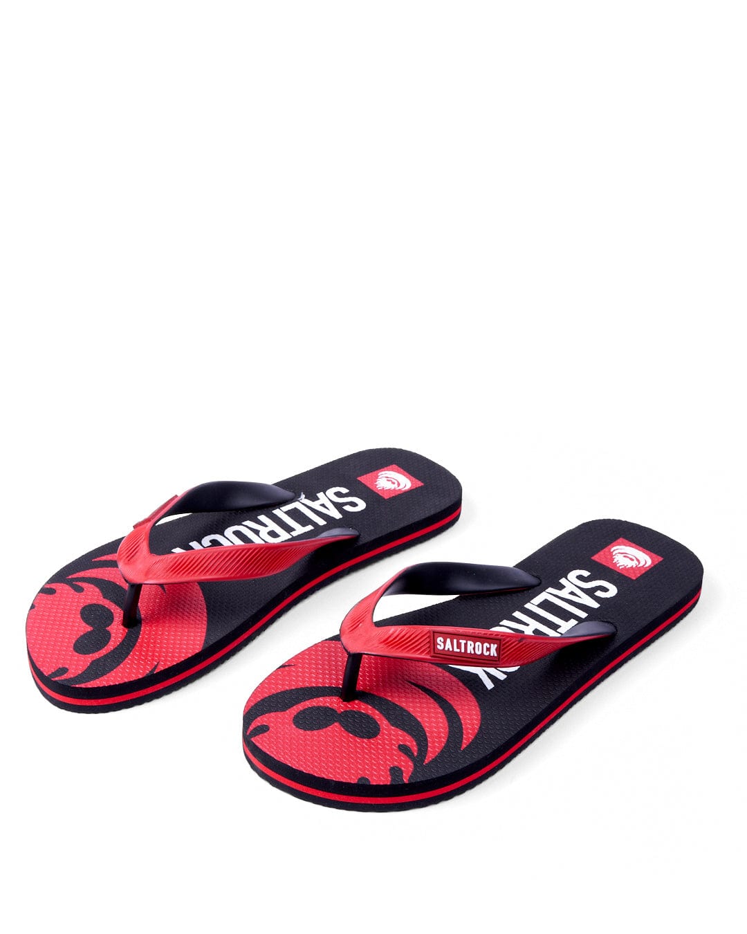 A pair of Saltrock Corp Flame flip-flops with red footbeds featuring large flamehead skulls, black straps, and the brand name in white letters.