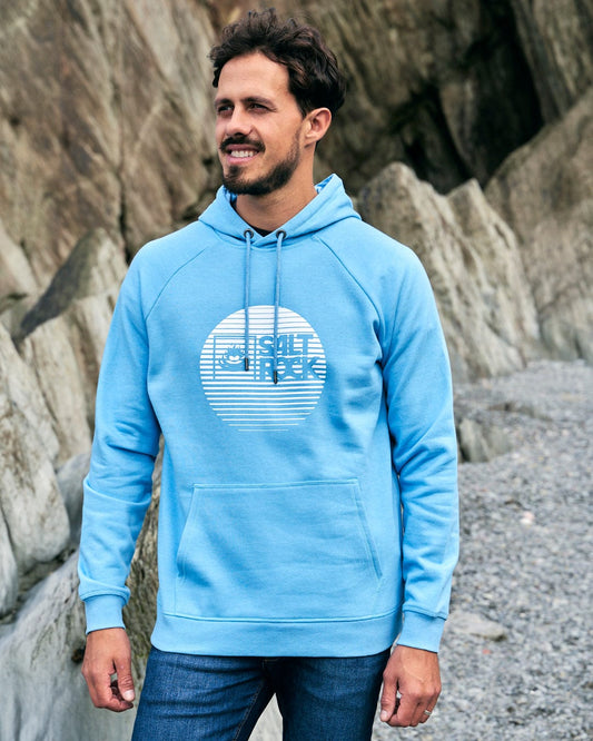 A man wearing a Corp Logo Fade - Mens Hoodie - Light Blue by Saltrock standing on rocks.