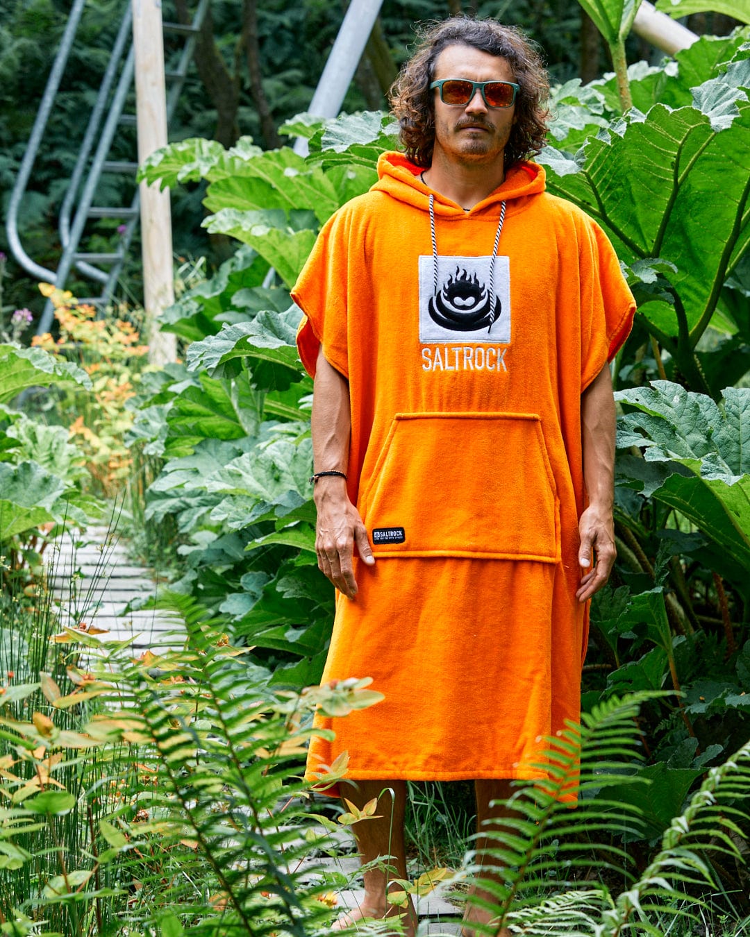 A person wearing the Saltrock Corp Changing Towel in orange, made from 100% cotton towelling with "SALTROCK" printed on it, stands on a pathway surrounded by lush green plants.