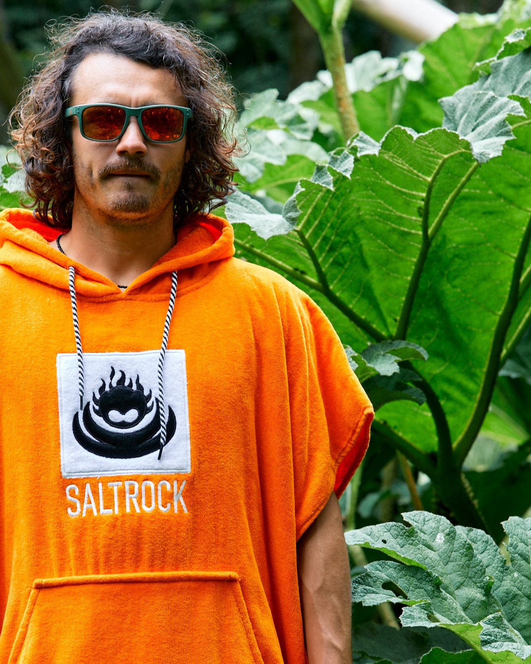 A person with curly hair and sunglasses stands outdoors wearing an orange Corp Changing Towel by Saltrock, made of 100% cotton towelling. Large green leaves are visible in the background.