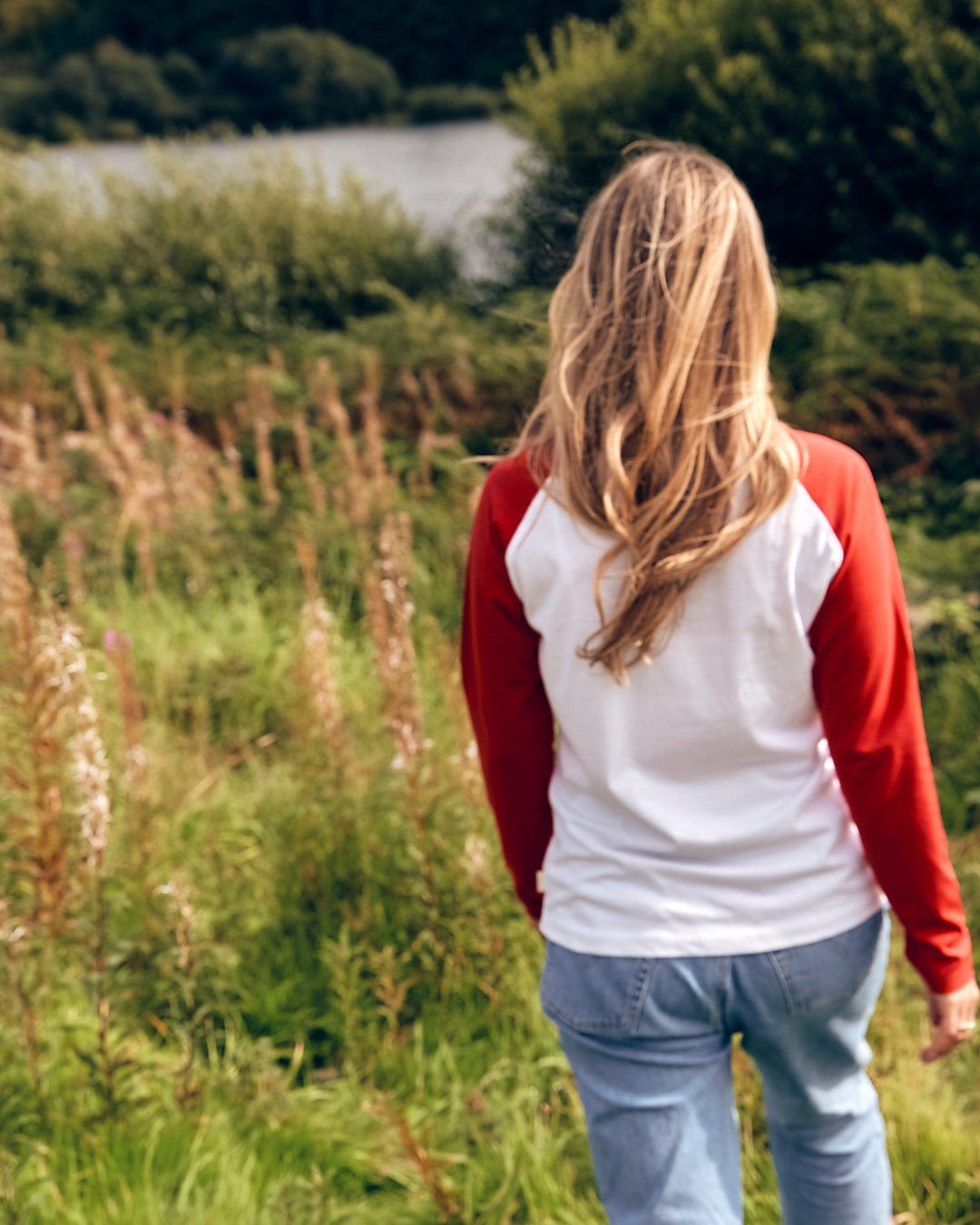 A woman walking through a field with a lake in the background wearing the Carolina - Womens Long Sleeve Raglan T-Shirt by Saltrock in Red/White.
