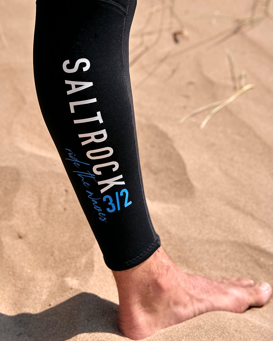 The legs of a person standing in the sand wearing a Saltrock Core - Mens 3/2 Full Wetsuit - Blue/Black.