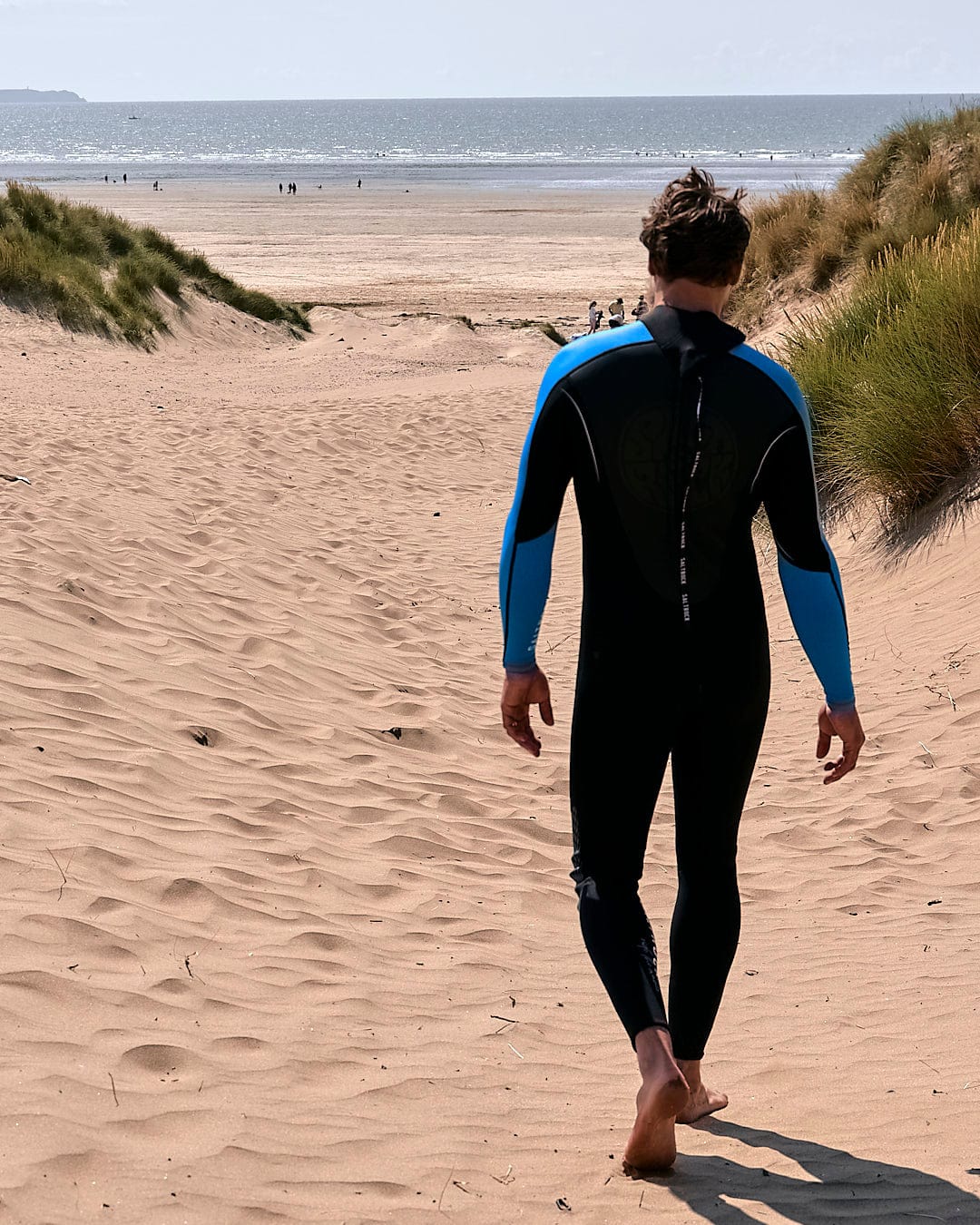 A man in a Saltrock Core - Mens 3/2 Full Wetsuit - Blue/Black walking down a beach.