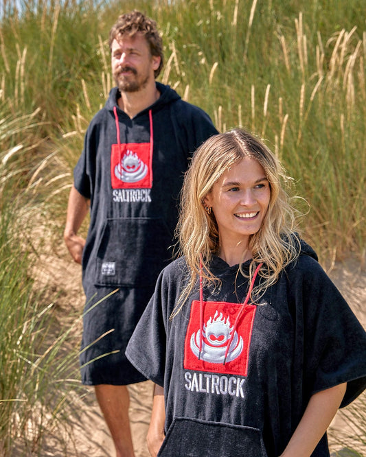 Two individuals wearing Saltrock branded Corp Changing Towel - Black, absorbent cotton towelling material clothing standing in a natural beach environment with tall grasses.