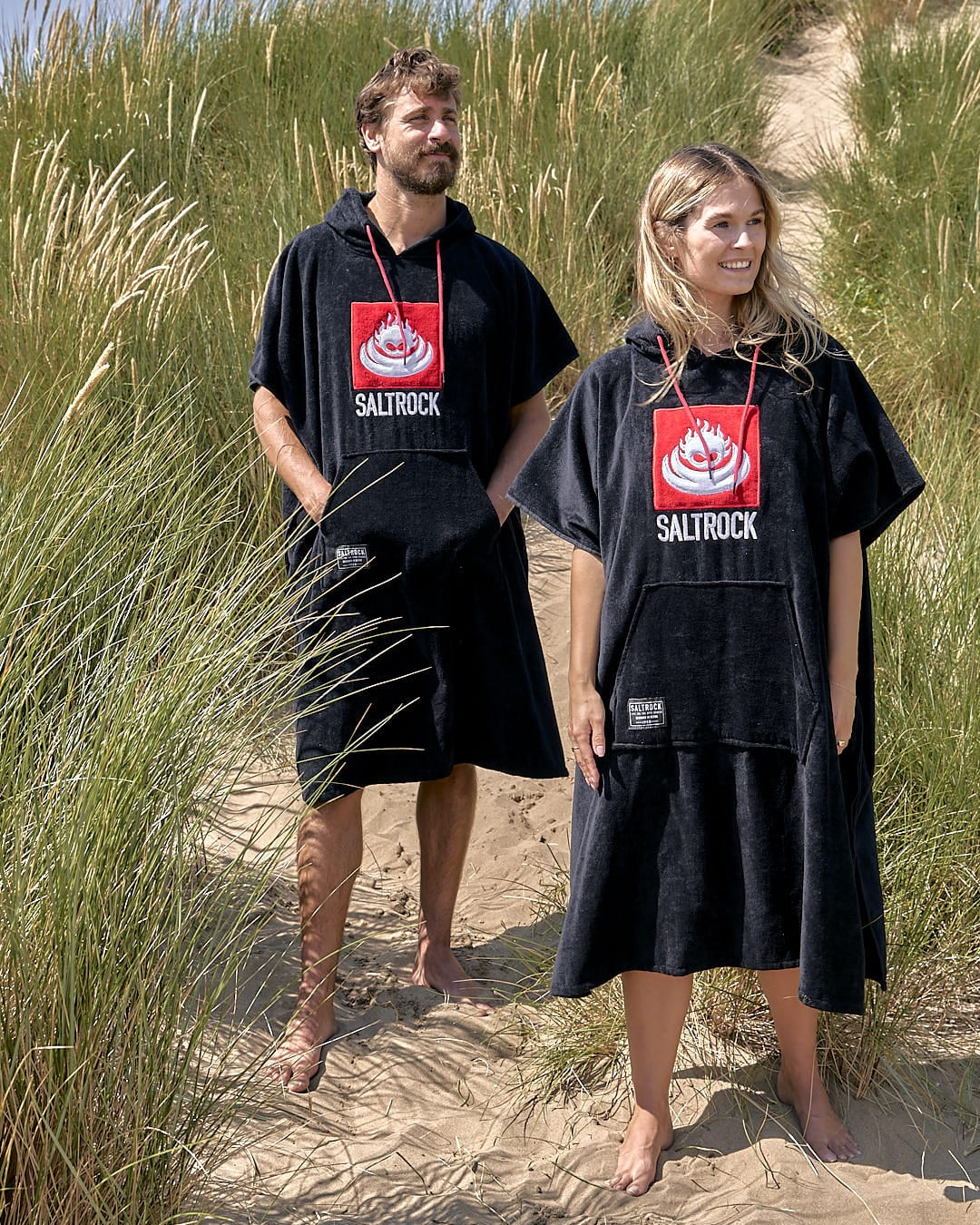 A man and a woman standing in the sand with Saltrock's Corp - Changing Towel - Black/Red.