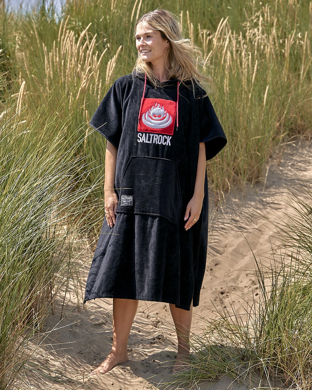 A woman wearing a Saltrock Corp - Changing Towel - Black/Red standing in the sand.