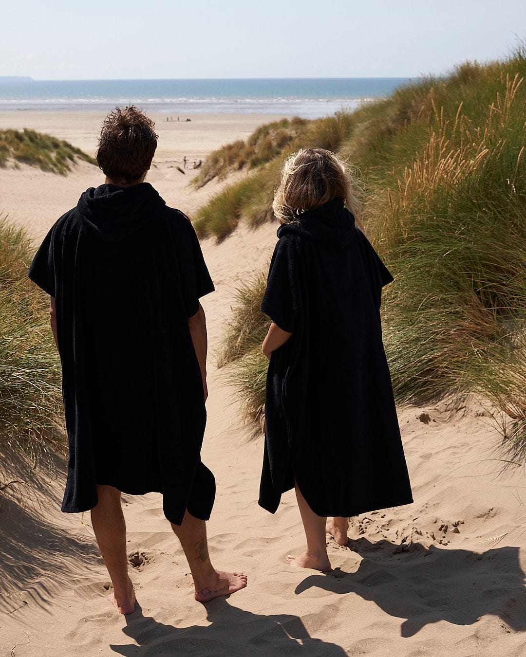 Two individuals wearing Saltrock Corp Changing Towel - Black robes walking on a sandy beach dune.