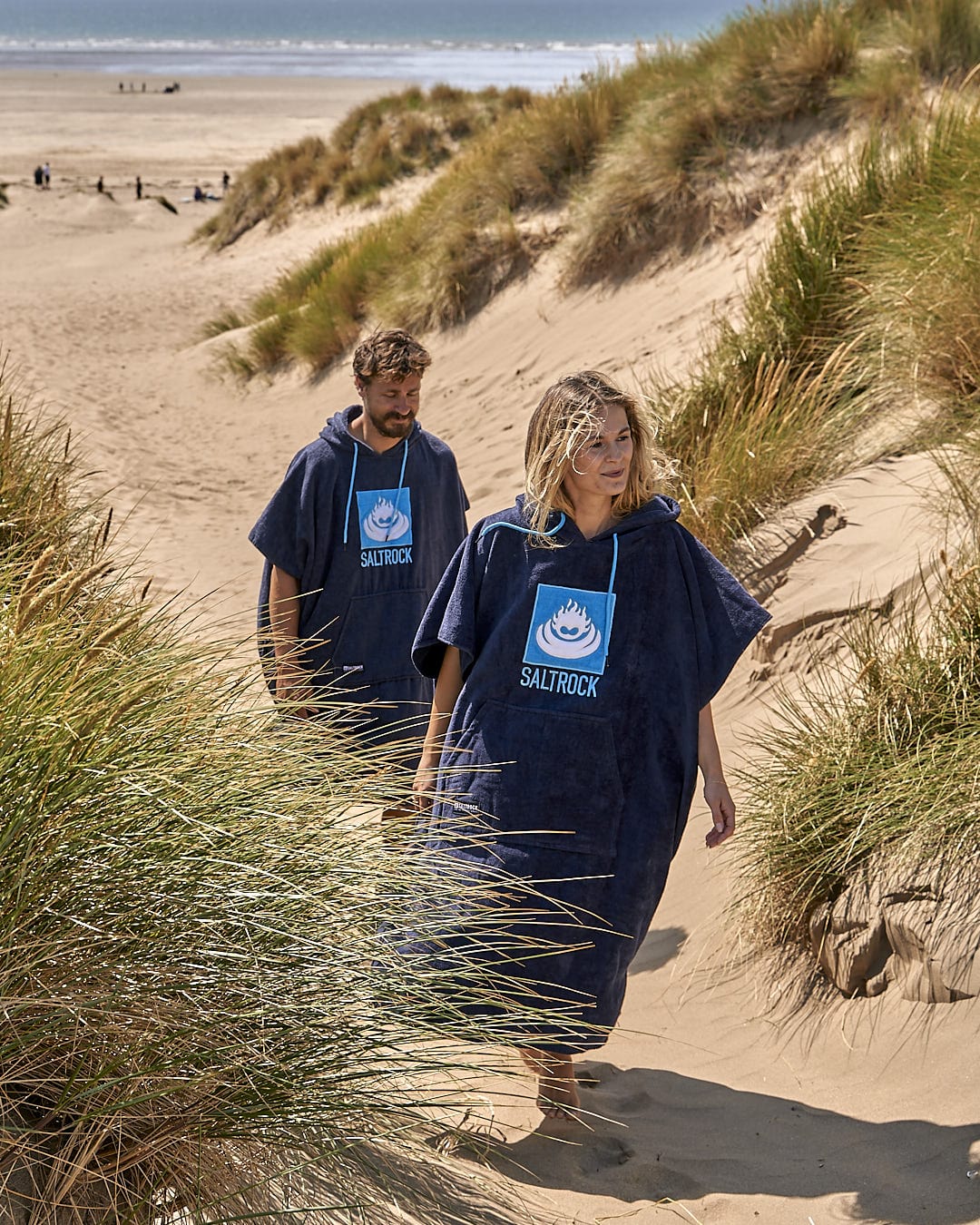 Two people walking through a sand dune on a beach while carrying the Saltrock Corp - Changing Towel - Blue.