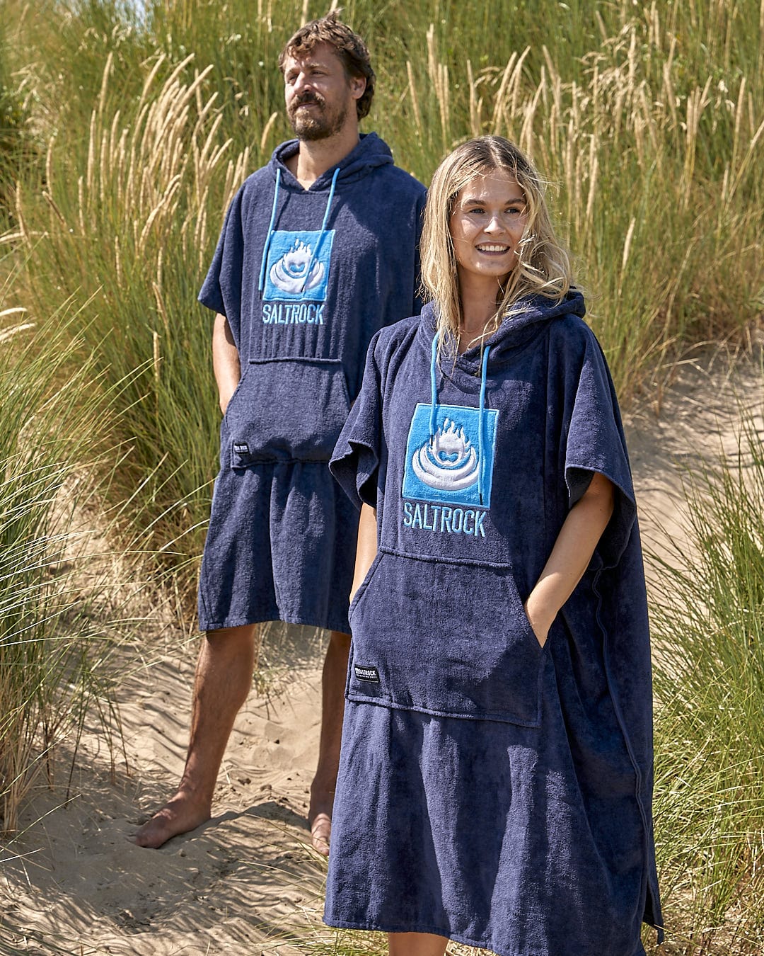 A man and woman standing in the sand wearing a Saltrock Corp - Changing Towel - Blue.