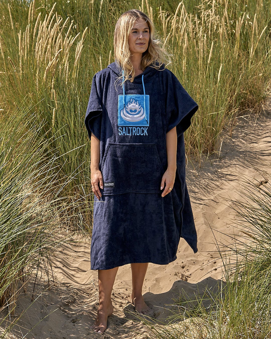 A woman standing in the sand wearing a Saltrock Corp - Changing Towel - Blue poncho.