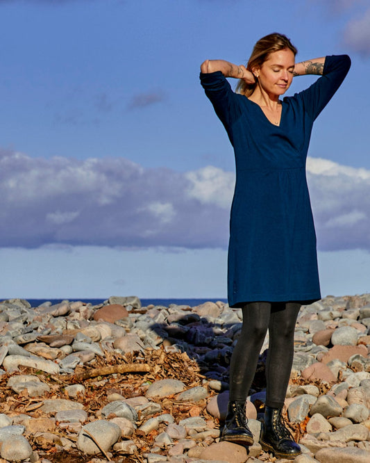 A fashion-conscious woman wearing a blue dress, Saltrock-branded Della - Womens Wrap Top Dress - Dark Blue, standing on a rocky beach.