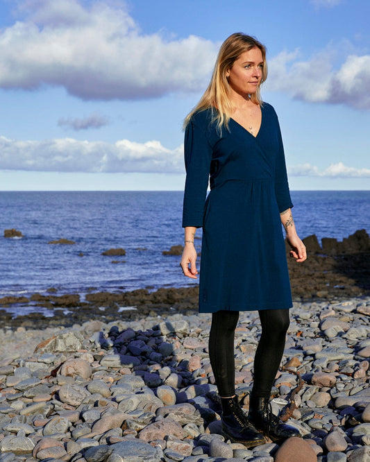 A fashion-conscious woman in a Saltrock Della - Womens Wrap Top Dress - Dark Blue standing on a rocky beach.