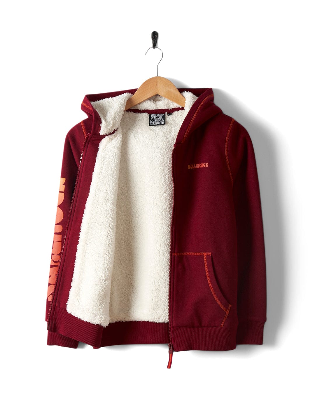 A wooden hanger showcases the Depeche Kids Sherpa Lined Hoodie in red, by Saltrock. It features a white fleece lining, bold sleeve branding, and a small chest logo.