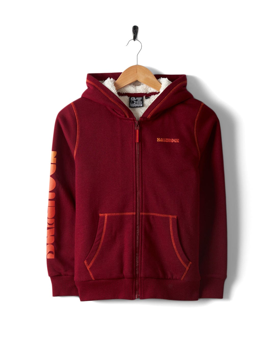A red Depeche Kids Sherpa Lined Hoodie by Saltrock, featuring a hood, front pockets, and "BARBOUR" in vibrant orange on the sleeve, hangs on a wooden hanger against a white background.