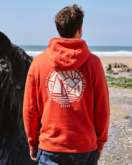 A man wearing a Saltrock Devon Sailaway - Mens Pop Hoodie - Red looking at the ocean.