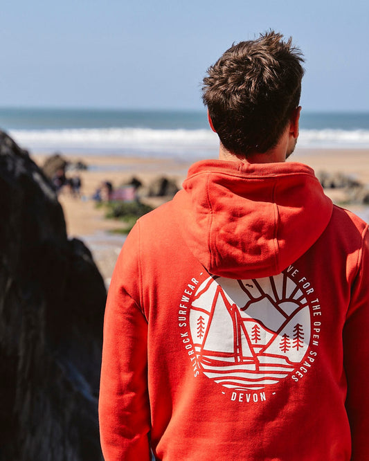 A man wearing a Saltrock Devon Sailaway - Mens Pop Hoodie - Red looking at the beach.