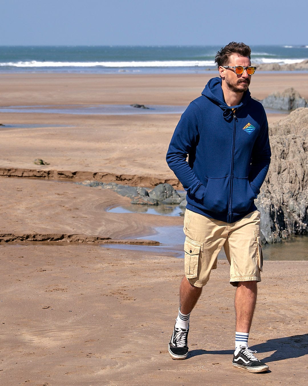A man wearing a Saltrock Diamond Scene - Mens Zip Hoodie - Blue and shorts walking on a beach.