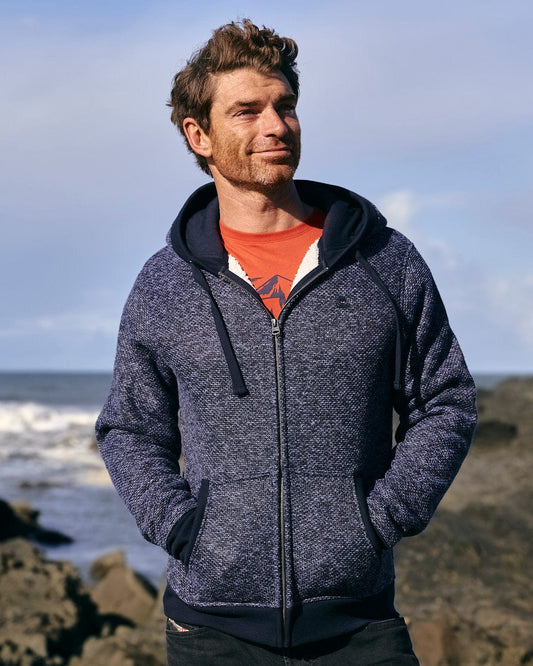 A man in a cozy blue Saltrock Dillan Lined Hoodie standing on rocks near the ocean.