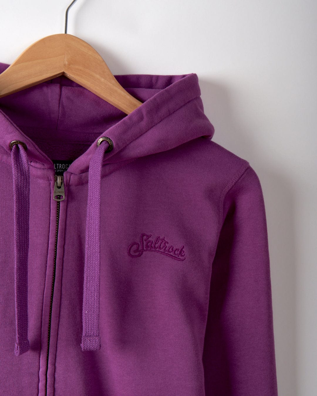 The Dip Dye Women's Zip Hoodie in purple, hanging on a wooden hanger against a white wall, showcases the Saltrock branding with an embroidered logo on the chest. Made from 100% cotton, it offers ultimate comfort and style.
