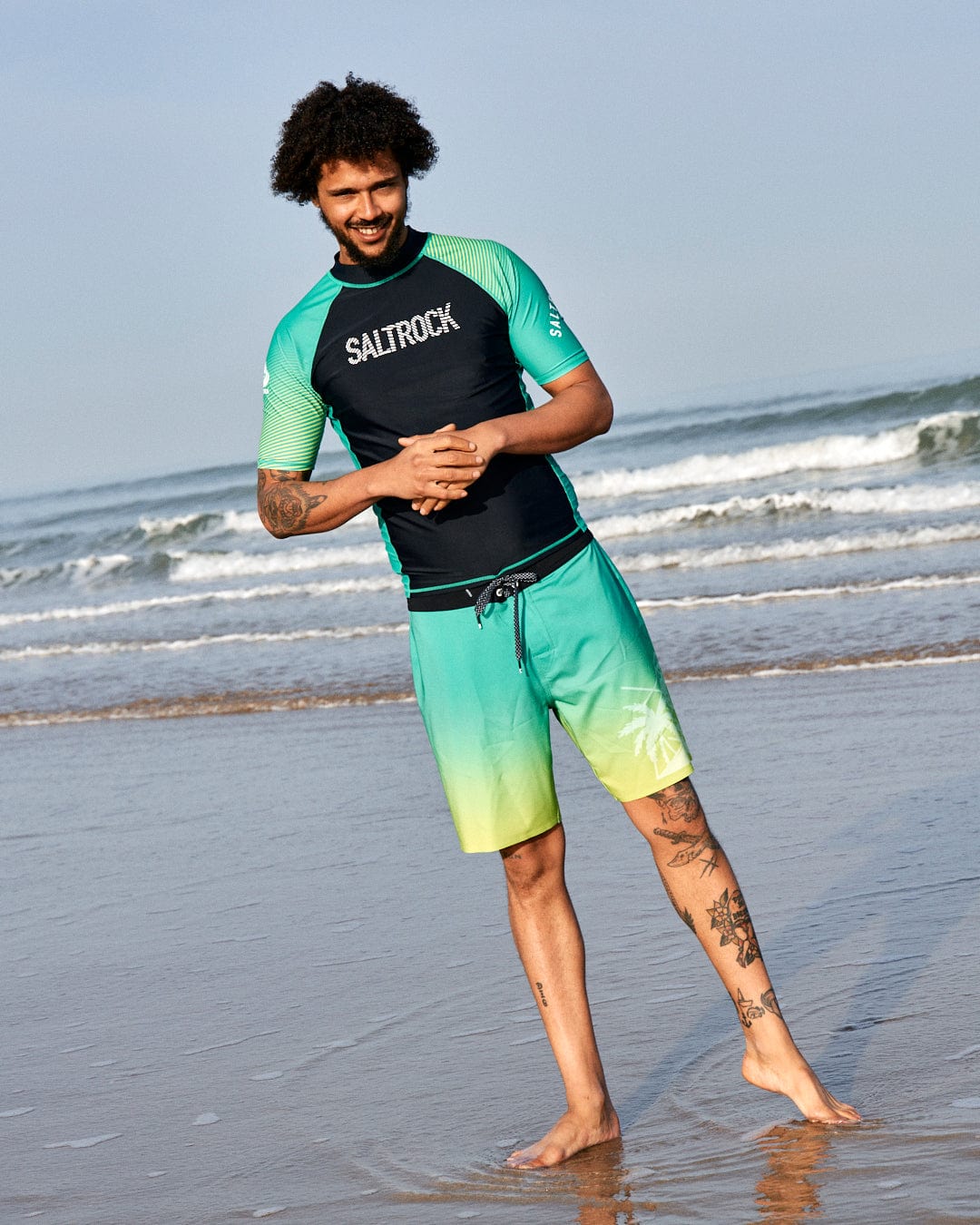 A man with curly hair stands on a beach, wearing a Saltrock DNA Wave Recycled Mens Short Sleeve Rashvest in Black/Green, and shorts, smiling towards the camera.