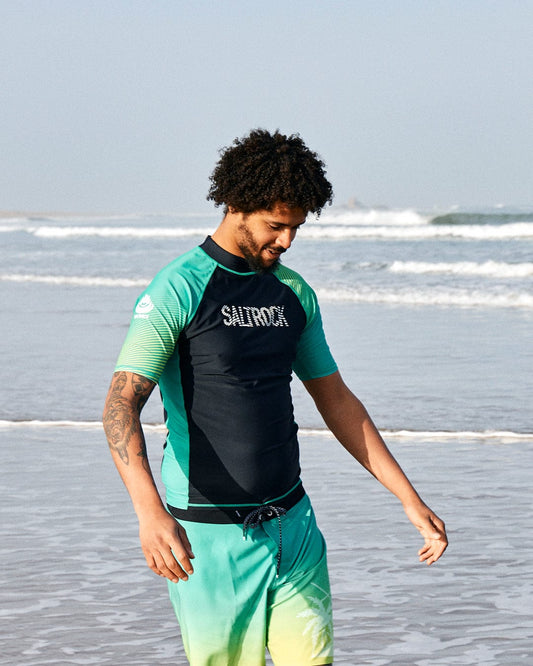 A man with curly hair wearing a Saltrock DNA Wave - Recycled Mens Short Sleeve Rashvest in Black/Green and Saltrock board shorts, both crafted from recycled polyester, stands on a beach, smiling down as he touches his waist.