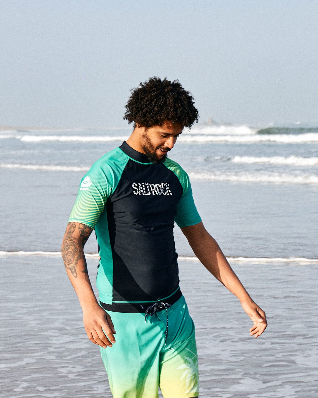 A man with curly hair wearing a Saltrock DNA Wave - Recycled Mens Short Sleeve Rashvest in Black/Green and Saltrock board shorts, both crafted from recycled polyester, stands on a beach, smiling down as he touches his waist.