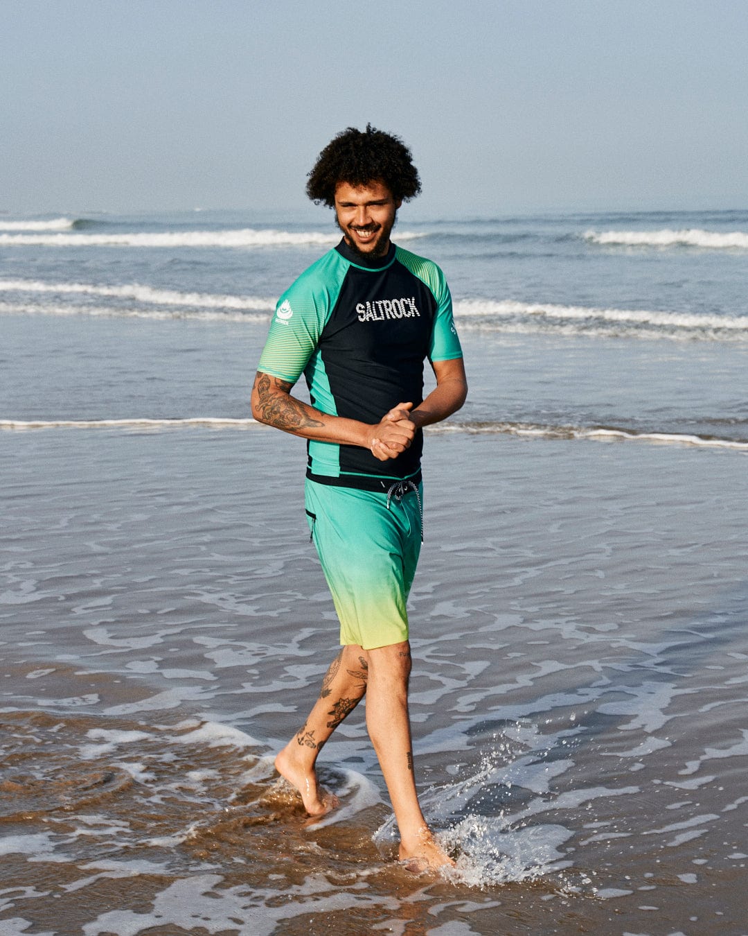 Man with curly hair in a Saltrock DNA Wave - Recycled Mens Short Sleeve Rashvest - Black/Green wetsuit, walking on a beach, smiling, with waves touching his feet.