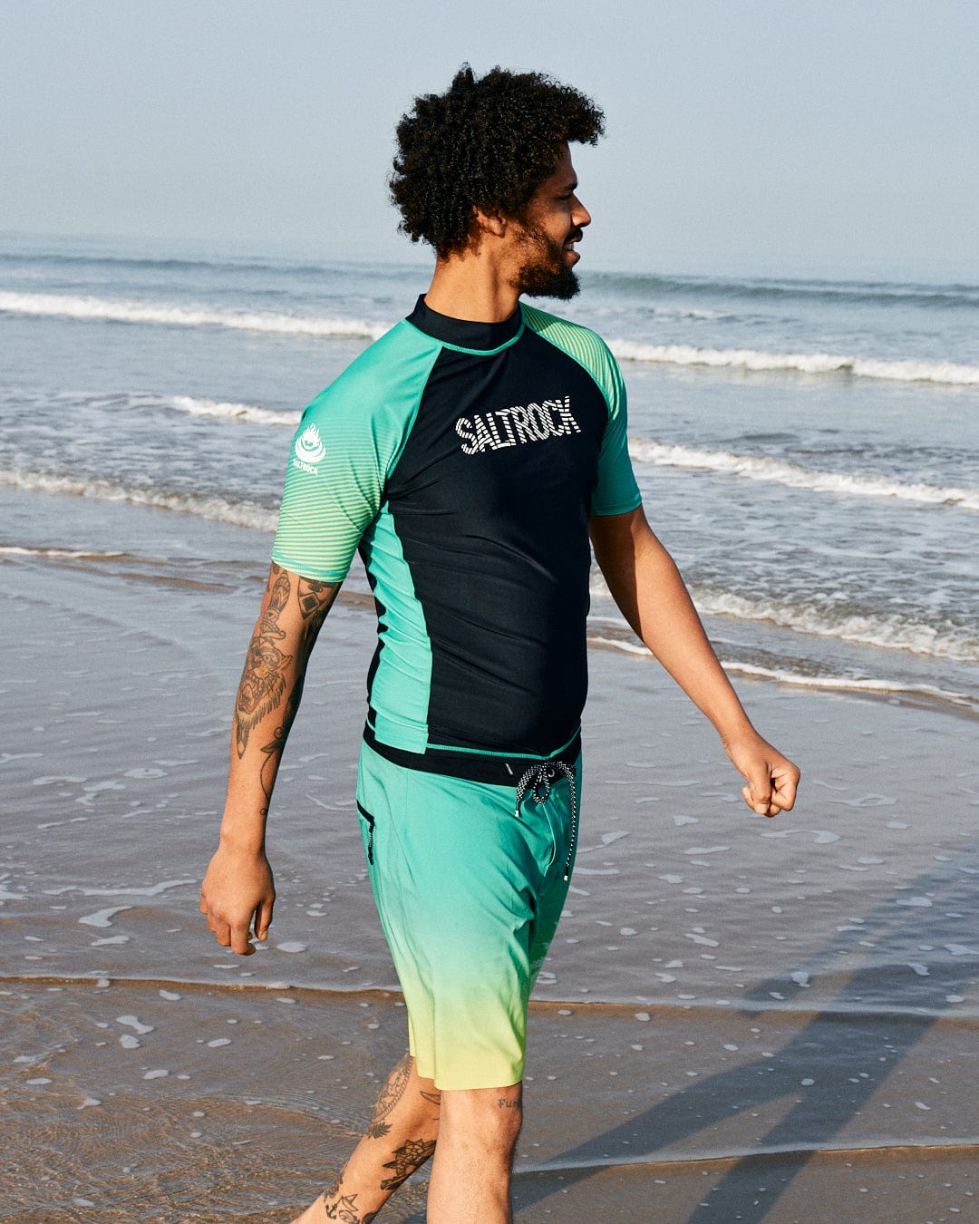 A man with curly hair, wearing a Saltrock DNA Wave Recycled Mens Short Sleeve Rashvest in Black/Green and teal shorts, walks along a sandy beach with the ocean in the background.