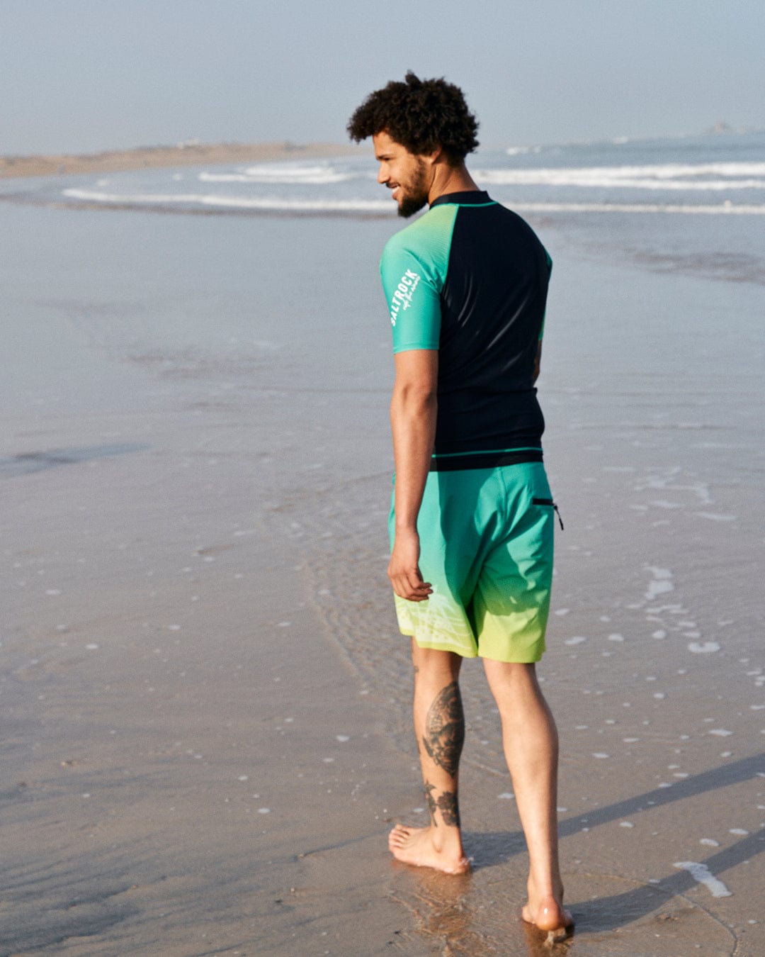A man with curly hair, wearing a Saltrock DNA Wave - Recycled Mens Short Sleeve Rashvest in Black/Green and board shorts, walks along a sandy beach, glancing over his shoulder.