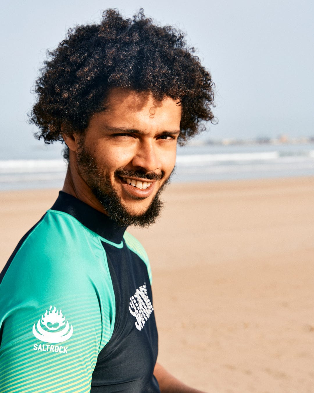 A man with curly hair wearing a green Saltrock DNA Wave Rashvest featuring UPF 50 protection smiles at the beach, with the ocean and sand visible in the background.