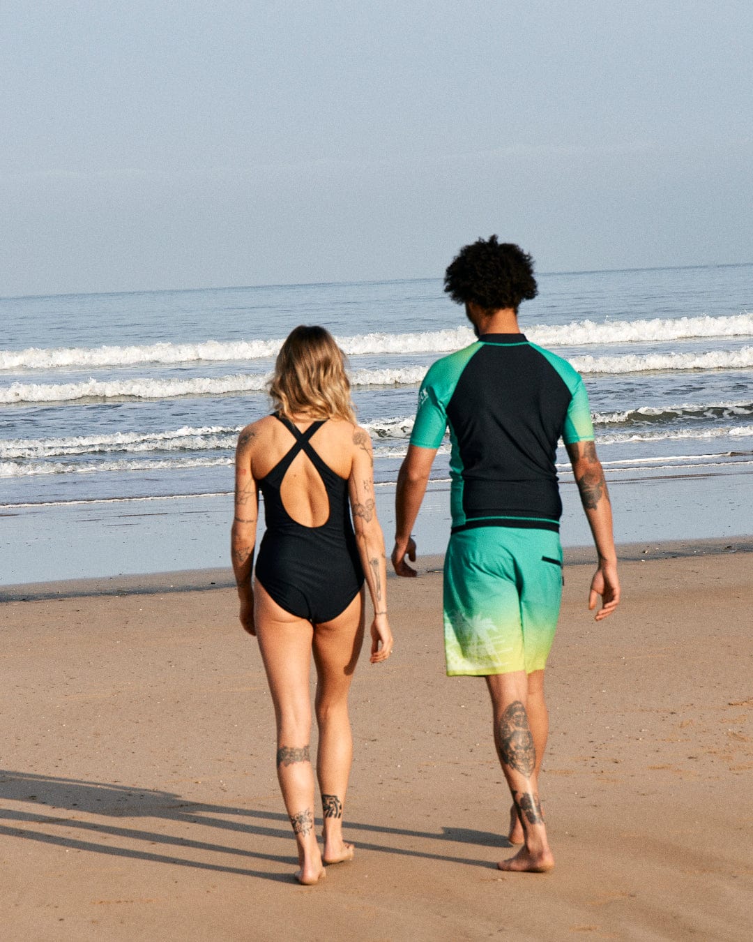 Two people walking on a beach, one in a Saltrock black swimsuit and the other in a Saltrock DNA Wave recycled mens short sleeve rashvest in black/green, both facing the ocean.