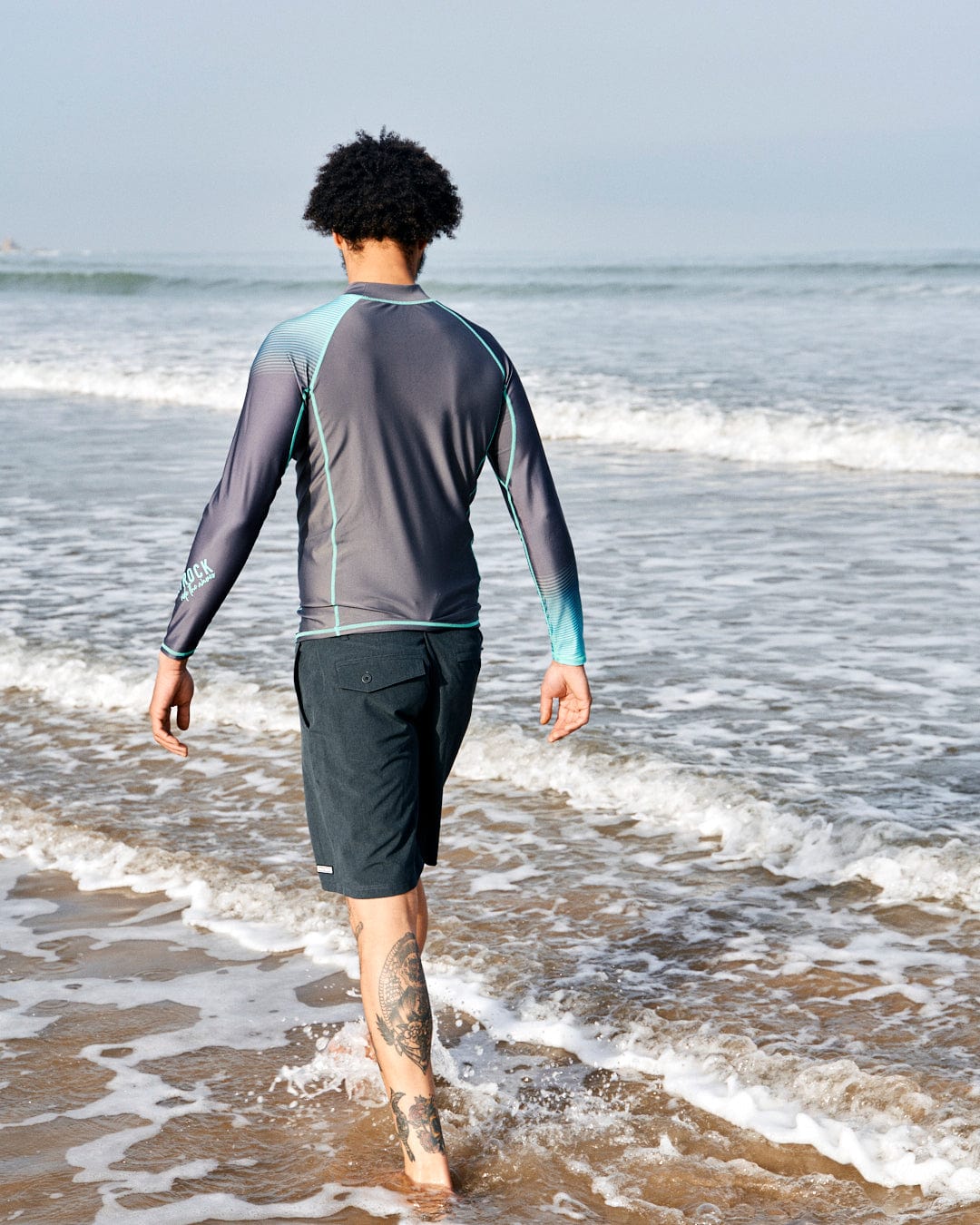 A man with curly hair walking into the sea, wearing a men's long-sleeved DNA Wave - Mens Long Sleeve Rashvest in Grey/Turquoise and shorts, and showing a tattoo on his left calf.
