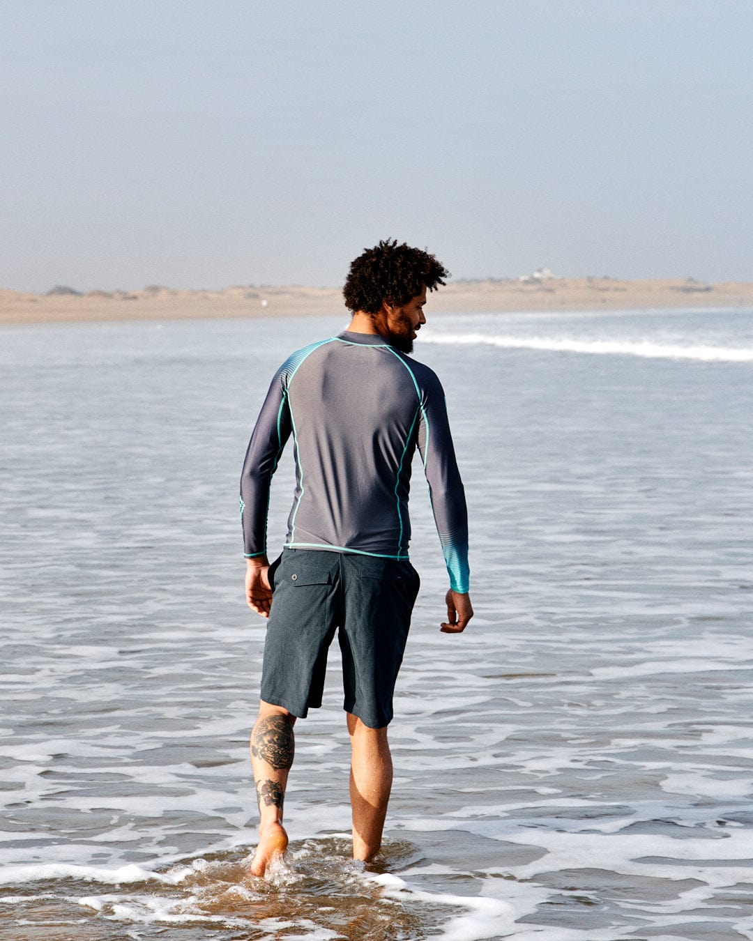 A man with curly hair and a tattoo on his calf walks along the shoreline, wearing a grey and teal Saltrock DNA Wave - Mens Long Sleeve Rashvest and dark shorts.
