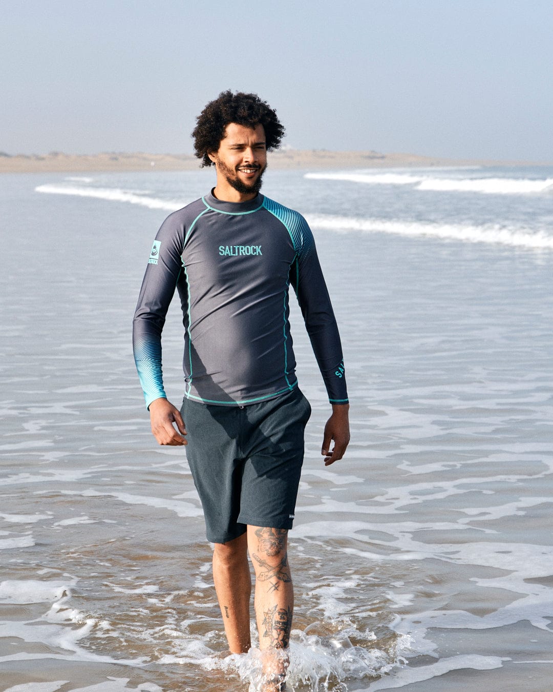 A man with curly hair wearing a Saltrock DNA Wave rash vest walks along a sandy beach with mild ocean waves at his feet.